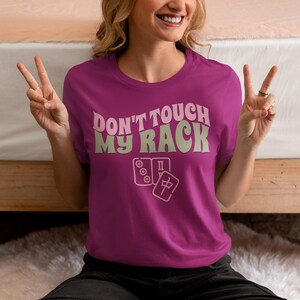 Może przedstawiać: R&oacute;żowy T-shirt z napisem "Don't touch my rack" w retro czcionce. Tekst jest w jasnozielonym kolorze. Pod tekstem znajdują się dwa kostki mahjong.