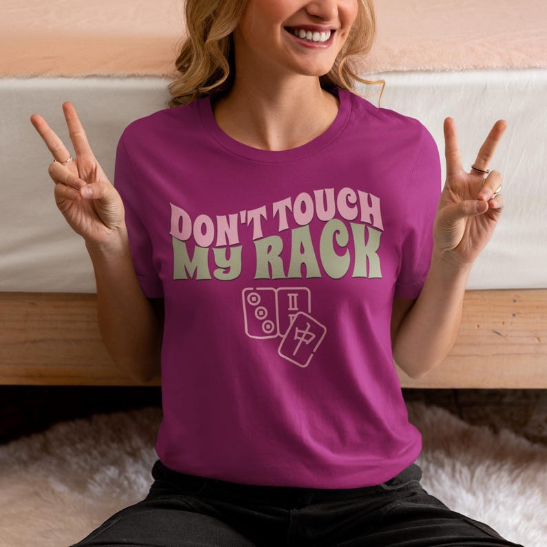 Może przedstawiać: R&oacute;żowy T-shirt z napisem "Don't touch my rack" w retro czcionce. Tekst jest w jasnozielonym kolorze. Pod tekstem znajdują się dwa kostki mahjong.