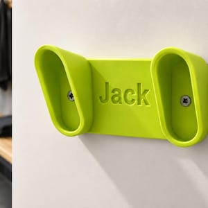 May include: A lime green wall-mounted shoe rack featuring two oval-shaped holders and the name Jack embossed in the centre. The unit is secured to the wall with two visible silver screws, providing a functional storage solution for footwear.