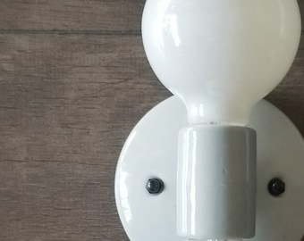 May include: White ceramic wall sconce with two white globe light bulbs. The sconce has a simple design with a cylindrical center piece and two round bases for the bulbs.