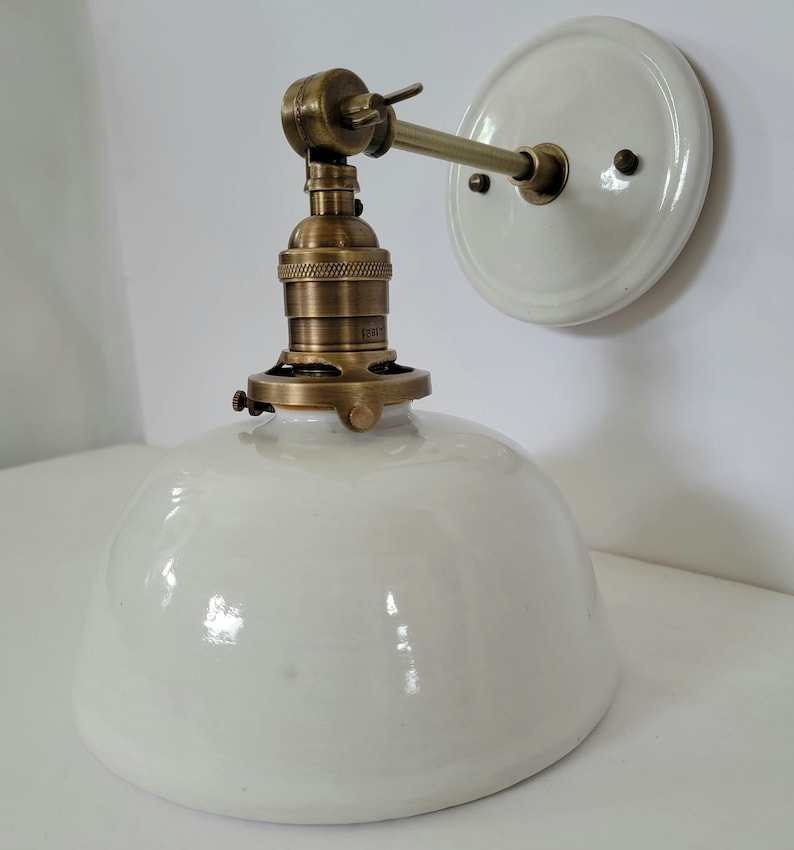 May include: White porcelain wall sconce with a brass socket and arm. The sconce has a white porcelain shade and a round back plate.