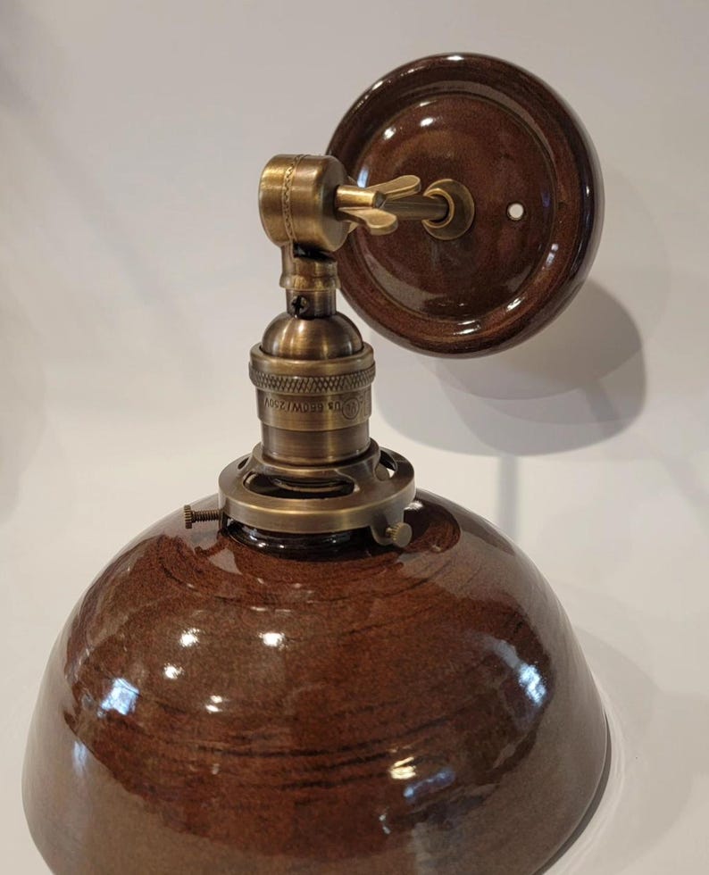 May include: A brown ceramic wall sconce with a brass socket and a brown ceramic shade. The sconce has a simple design and is perfect for adding a touch of rustic charm to any room.