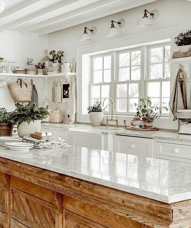 May include: A white kitchen with a large window, white cabinets, and a wooden island with a white countertop. The island has a farmhouse style with a chevron pattern on the front. There are plants on the windowsill and shelves.