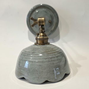May include: A ceramic wall sconce with a scalloped shade in a muted grey-blue hue. The fixture has a brass-coloured socket and a matching circular backplate. The design is a blend of rustic and industrial styles.