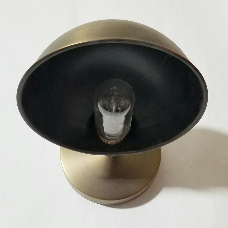 Peut inclure: Une applique murale en laiton avec un abat-jour noir et une ampoule transparente. L'applique a un design simple et moderne.