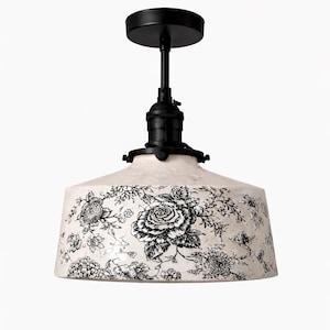 May include: A pendant light fixture with a black metal frame and a cream-colored shade. The shade is decorated with a black floral pattern. The light fixture is designed to be mounted on a ceiling.