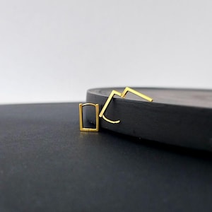 Può includere: Orecchini geometrici color oro. Uno è un cerchio rettangolare, l'altro ha un design a zig-zag. Gli orecchini sono esposti su una superficie grigio scuro su sfondo bianco. I gioielli sono semplici e moderni.