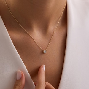 Op de afbeelding: Een delicate gouden ketting met een kleine, vierkante hanger met een enkele, heldere edelsteen. De hanger hangt aan een dunne gouden ketting. De ketting wordt gedragen over een witte top.