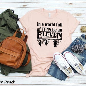 以下が含まれることがあります： ピンクのTシャツに「In a world full of TENS be an ELEVEN」という黒い文字が書かれています。文字はレトロなフォントで、黒い枠で囲まれています。文字の上に、自転車に乗った人の黒いシルエットが4つあります。