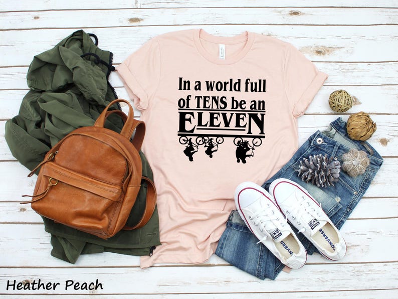 以下が含まれることがあります： ピンクのTシャツに「In a world full of TENS be an ELEVEN」という黒い文字が書かれています。文字はレトロなフォントで、黒い枠で囲まれています。文字の上に、自転車に乗った人の黒いシルエットが4つあります。
