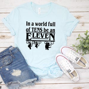 以下が含まれることがあります： 薄い青色のTシャツに、黒い文字で「In a world full of TENS be an ELEVEN」と書いてあります。テキストは太字で、黒い枠で囲まれています。テキストの上に、自転車に乗った人の黒いシルエットが3つあります。