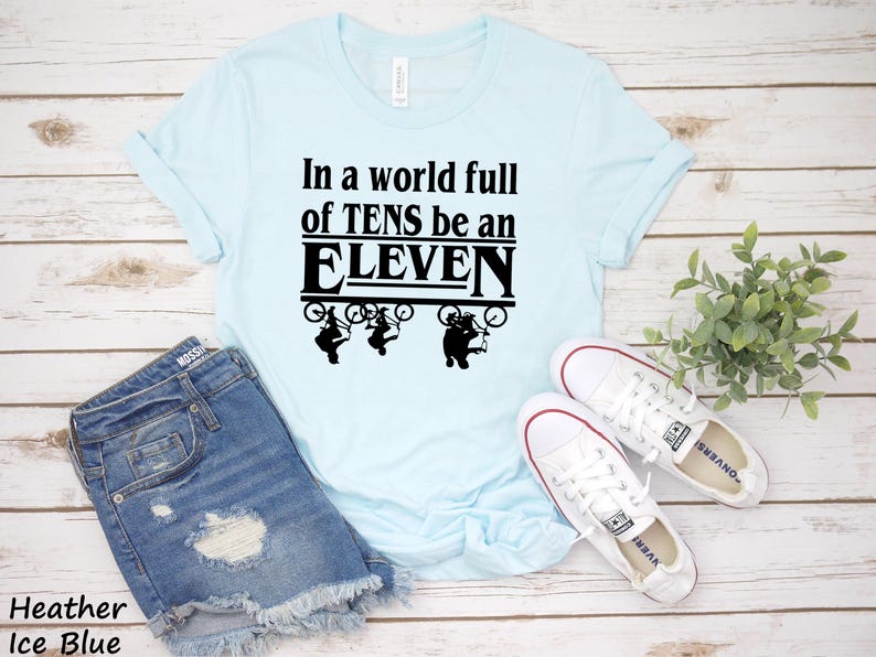 以下が含まれることがあります： 薄い青色のTシャツに、黒い文字で「In a world full of TENS be an ELEVEN」と書いてあります。テキストは太字で、黒い枠で囲まれています。テキストの上に、自転車に乗った人の黒いシルエットが3つあります。