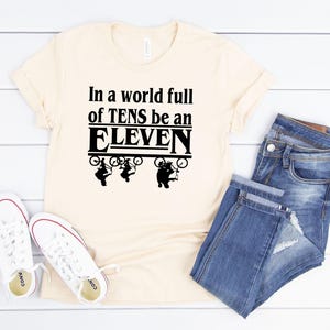 以下が含まれることがあります： ソフトクリーム色のTシャツに、黒い文字で「In a world full of TENS be an ELEVEN」と書いてあります。テキストの周りを、自転車に乗った4人の黒いシルエットが囲んでいます。