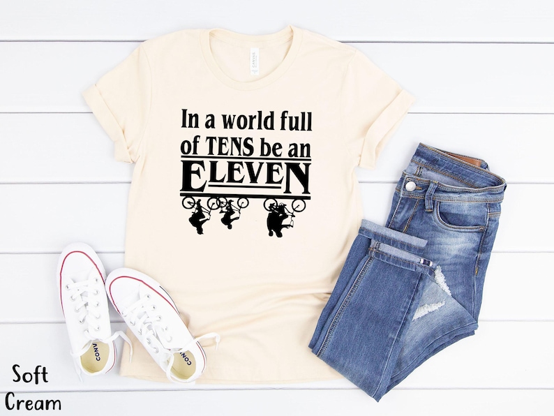 以下が含まれることがあります： ソフトクリーム色のTシャツに、黒い文字で「In a world full of TENS be an ELEVEN」と書いてあります。テキストの周りを、自転車に乗った4人の黒いシルエットが囲んでいます。