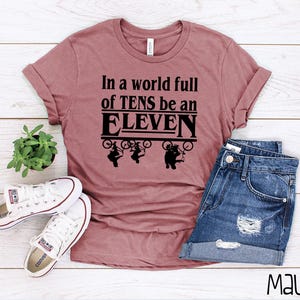 以下が含まれることがあります： マゼンタ色のTシャツに、黒い文字で「In a world full of TENS be an ELEVEN」と書いてあります。テキストは、自転車に乗った4人の人物のグラフィックで囲まれています。