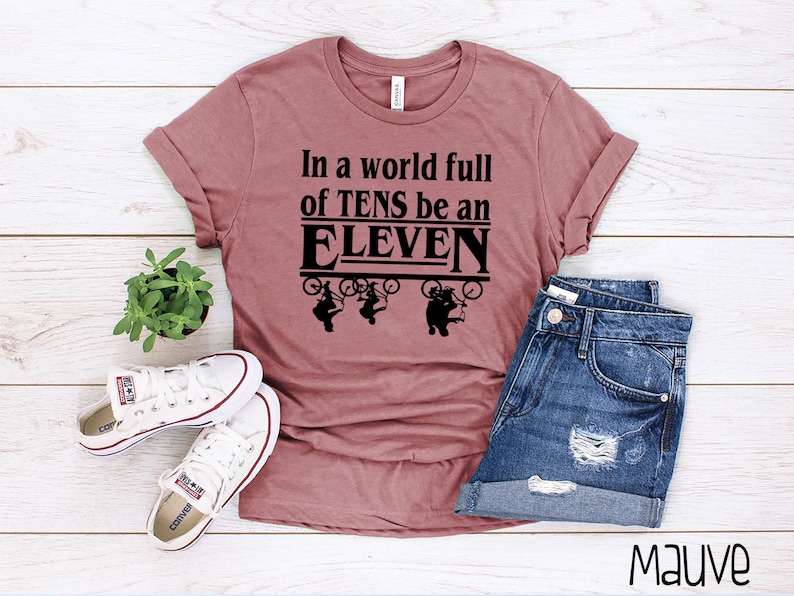 以下が含まれることがあります： マゼンタ色のTシャツに、黒い文字で「In a world full of TENS be an ELEVEN」と書いてあります。テキストは、自転車に乗った4人の人物のグラフィックで囲まれています。