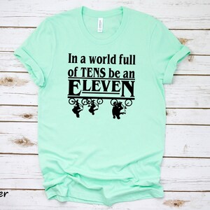 以下が含まれることがあります： 薄い緑色のTシャツに、黒い文字で「In a world full of TENS be an ELEVEN」と書かれています。テキストは、自転車に乗った3人の人物の黒いグラフィックで囲まれています。