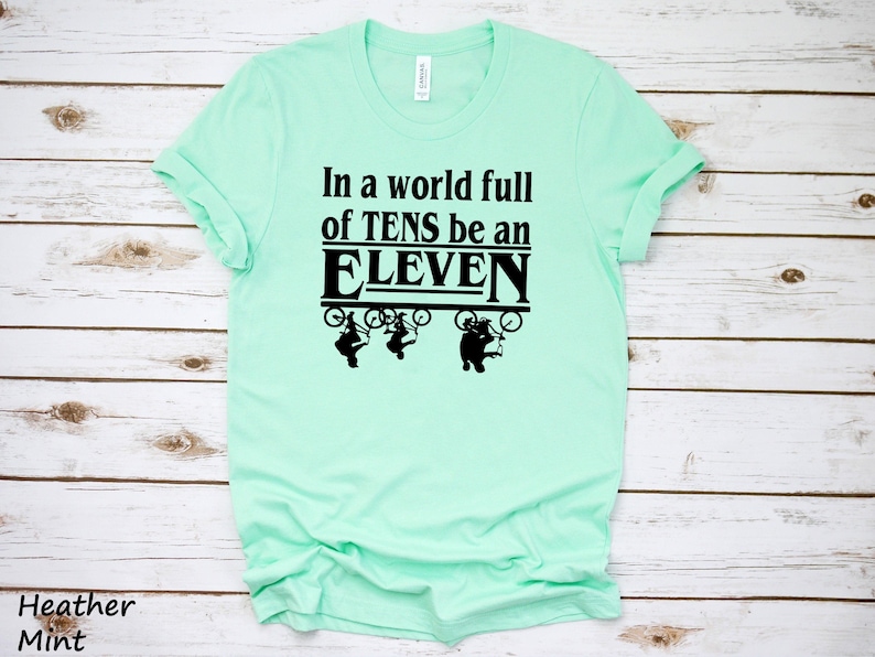 以下が含まれることがあります： 薄い緑色のTシャツに、黒い文字で「In a world full of TENS be an ELEVEN」と書かれています。テキストは、自転車に乗った3人の人物の黒いグラフィックで囲まれています。
