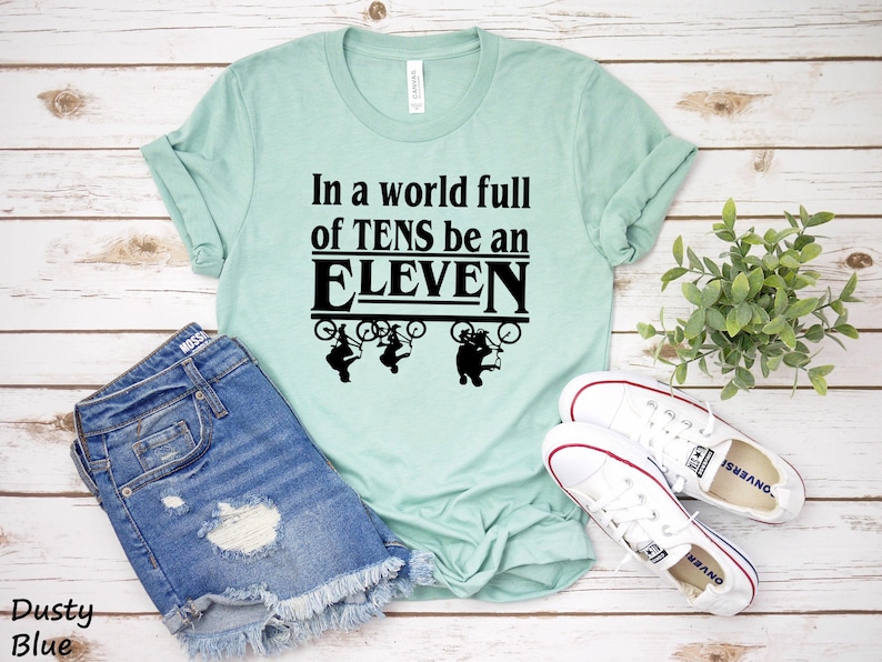 以下が含まれることがあります： 薄い青色のTシャツに、黒い文字で「In a world full of TENS be an ELEVEN」と書いてあります。テキストは太字で、黒い枠で囲まれています。テキストの上に、自転車に乗った人の黒いシルエットが3つあります。