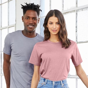 以下が含まれることがあります： 窓の前に立っている2人の人物。一人は薄いグレーの無地Tシャツを着ており、もう一人はくすんだピンクの無地Tシャツを着ています。