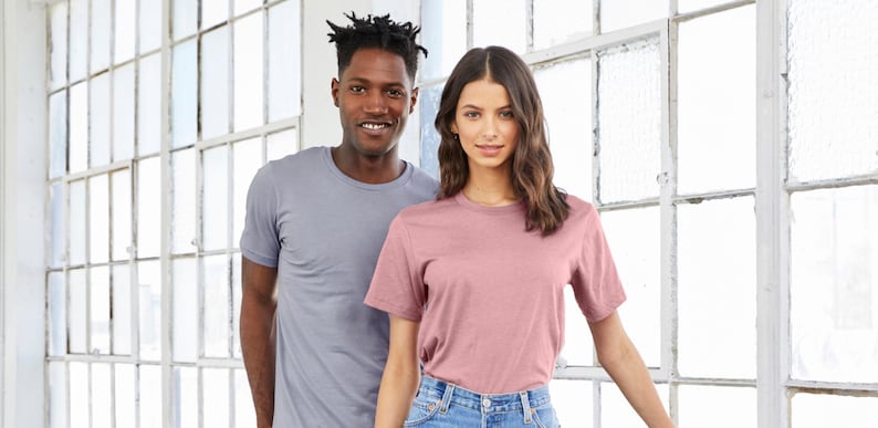 以下が含まれることがあります： 窓の前に立っている2人の人物。一人は薄いグレーの無地Tシャツを着ており、もう一人はくすんだピンクの無地Tシャツを着ています。
