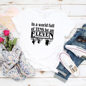 以下が含まれることがあります： 黒い文字で「In a world full of TENS be an ELEVEN」と書かれた白いTシャツ。テキストは黒い枠で囲まれています。テキストの上に、自転車に乗った人の黒いシルエットが4つあります。