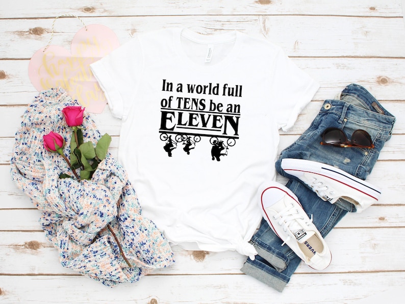 以下が含まれることがあります： 黒い文字で「In a world full of TENS be an ELEVEN」と書かれた白いTシャツ。テキストは黒い枠で囲まれています。テキストの上に、自転車に乗った人の黒いシルエットが4つあります。