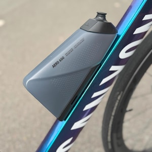 Puede incluir: Botella de agua Aero 650 gris, montada en un cuadro de bicicleta. La botella tiene una tapa gris oscuro y un dise&ntilde;o aerodin&aacute;mico. El cuadro de la bicicleta es oscuro con detalles iridiscentes azules y morados. El texto "AERO 650" est&aacute; impreso en la botella.