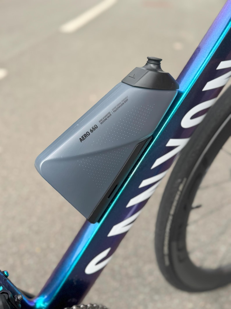 Puede incluir: Botella de agua Aero 650 gris, montada en un cuadro de bicicleta. La botella tiene una tapa gris oscuro y un dise&ntilde;o aerodin&aacute;mico. El cuadro de la bicicleta es oscuro con detalles iridiscentes azules y morados. El texto "AERO 650" est&aacute; impreso en la botella.