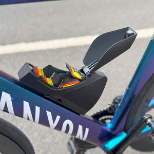 Puede incluir: Una caja de almacenamiento negra sujeta a un cuadro de bicicleta, parcialmente abierta para revelar paquetes coloridos. El cuadro de la bicicleta es un degradado de azul, morado y turquesa, con la marca "CANYON" en blanco.