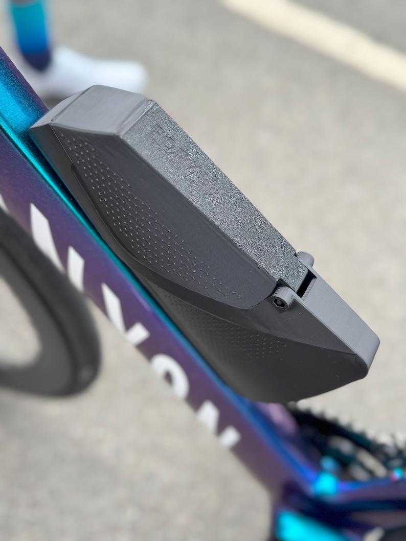 Puede incluir: Primer plano de una caja de almacenamiento gris montada en el cuadro de una bicicleta. La caja est&aacute; sujeta a una bicicleta con una combinaci&oacute;n de colores azul, morado y blanco. El texto "FOR K" es visible en la caja.
