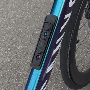Puede incluir: Accesorio de bicicleta negro montado en un cuadro de color turquesa y morado. El accesorio tiene dos botones circulares y una forma rectangular. El cuadro de la bicicleta tiene letras blancas y un neum&aacute;tico negro.