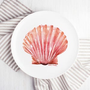 May include: A white ceramic plate featuring a watercolor painting of a pink and red seashell. The seashell design has vertical lines and a textured appearance. The plate is resting on a striped gray and white cloth.