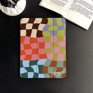 Könnte beinhalten: Eine farbenfrohe Tablet-Hülle mit einem wellenförmigen Schachbrettmuster in Braun, Orange, Rosa, Blau und Grün. Die Hülle hat ein weiches, geschwungenes Design und trägt den handgeschriebenen Namen "Anny" mit einem Herzsymbol.