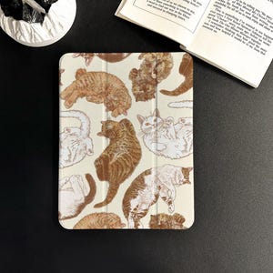 Puede incluir: Una funda de tableta de color crema con un patrón repetido de ilustraciones de gatos marrones y blancos en varias poses. La funda está abierta, revelando el interior. El diseño es de estilo caprichoso y dibujado a mano.