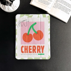 Op de afbeelding: Een iPad-hoes met een wit en groen patroon en een roze vierkant in het midden. Het vierkant bevat een tekening van twee rode kersen met groene stelen. De tekst "CHERRY" staat in rode letters onder de kersen. De tekst "DON'T YOU CALL HIM BABY - FINE LINE" staat in zwarte letters onder het woord "CHERRY". De tekst "Harry" is met zwarte inkt in de linkerbovenhoek van het hoes geschreven.