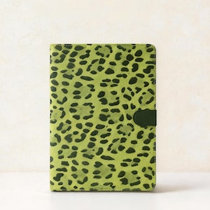 Puede incluir: Un diario con estampado de leopardo verde y negro con un cierre verde oscuro. La cubierta presenta un fondo verde vibrante con manchas verde oscuro. El diario es rectangular y parece estar hecho de cuero o un material similar.