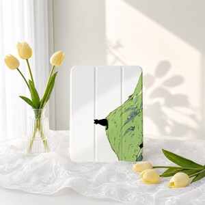 Puede incluir: Una funda para tableta blanca con una ilustración gráfica de un pequeño gato negro posado sobre una colina verde texturizada. La funda tiene un diseño de tres pliegues con líneas verticales visibles para proteger el dispositivo.