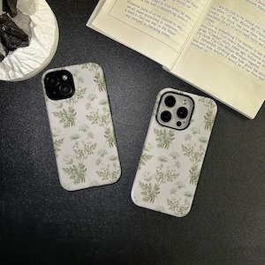 May include: Two white phone cases with a delicate floral pattern of green leaves and small white flowers. The cases are on a dark surface, with a book and a decorative item in the background.