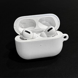 May include: White wireless earbuds in an open white charging case. The case has a small loop on the side. The earbuds are visible inside the case, ready for use. The background is black.