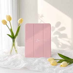 May include: A light pink tablet case with a tri-fold design and the name Lucie written in white cursive script across the centre. The case features a smooth, matte finish and is positioned upright, displaying a clean and simple aesthetic.