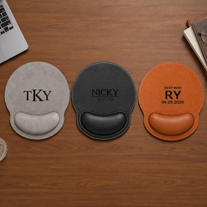 Può includere: Tre tappetini per mouse circolari con poggiapolsi in grigio, nero e arancione. Il tappetino grigio presenta il testo TKY. Quello nero mostra NICKY e 04.05.2026. Quello arancione mostra BEST MAN, RY e 04.05.2026. Ogni tappetino ha una superficie liscia.