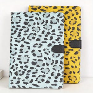 Puede incluir: Dos fundas para tablet con estampado animal. Una es azul claro con manchas negras, la otra es amarilla con manchas negras. Ambas tienen un cierre magnético de cuero negro.