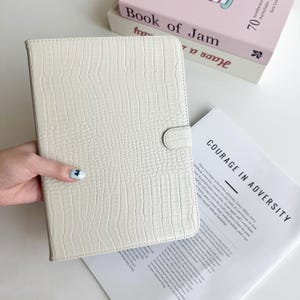 Può includere: Custodia per tablet color crema con texture a pelle di coccodrillo e chiusura a scatto. Il testo "COURAGE IN ADVERSITY" è visibile su una pagina accanto alla custodia.