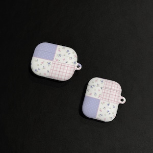 Puede incluir: Dos fundas blancas para AirPods con diseño de patchwork. Cada funda presenta secciones de lunares lavanda, cuadros rosas y estampados florales. Las fundas tienen un pequeño lazo para sujetar un cordón y están sobre un fondo negro.