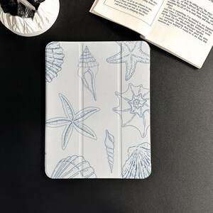 Puede incluir: Una funda blanca para tableta con un diseño de conchas marinas y estrellas de mar azules. El diseño incluye varias ilustraciones de conchas marinas, como conchas y estrellas de mar. La funda se muestra sobre una superficie negra con un libro y un objeto decorativo blanco en el fondo.