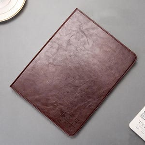 May include: A brown leather-bound folio with a textured surface. The folio has a stitched border and a debossed logo in the lower right corner. The folio is rectangular and appears to be closed.