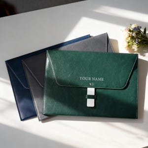 May include: Three leather document holders in navy, grey, and green. The green folder has the text "YOUR NAME". Each has a flap closure with white stitching and a white clasp. The folders are on a white surface.