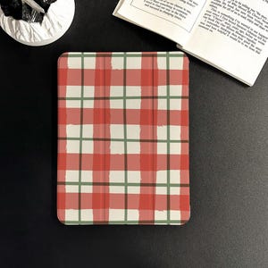 May include: A tablet case with a red, green, and white plaid pattern. The case is rectangular and has rounded corners. The design is reminiscent of a picnic blanket. The case is shown on a black surface with a book and a white object in the background.
