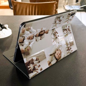 Puede incluir: Una funda negra para tableta con un dise&ntilde;o de collage de fotos. El collage presenta m&uacute;ltiples fotos familiares en una variedad de tama&ntilde;os y orientaciones. La funda est&aacute; abierta y apoyada, mostrando las im&aacute;genes. El fondo es una superficie oscura.